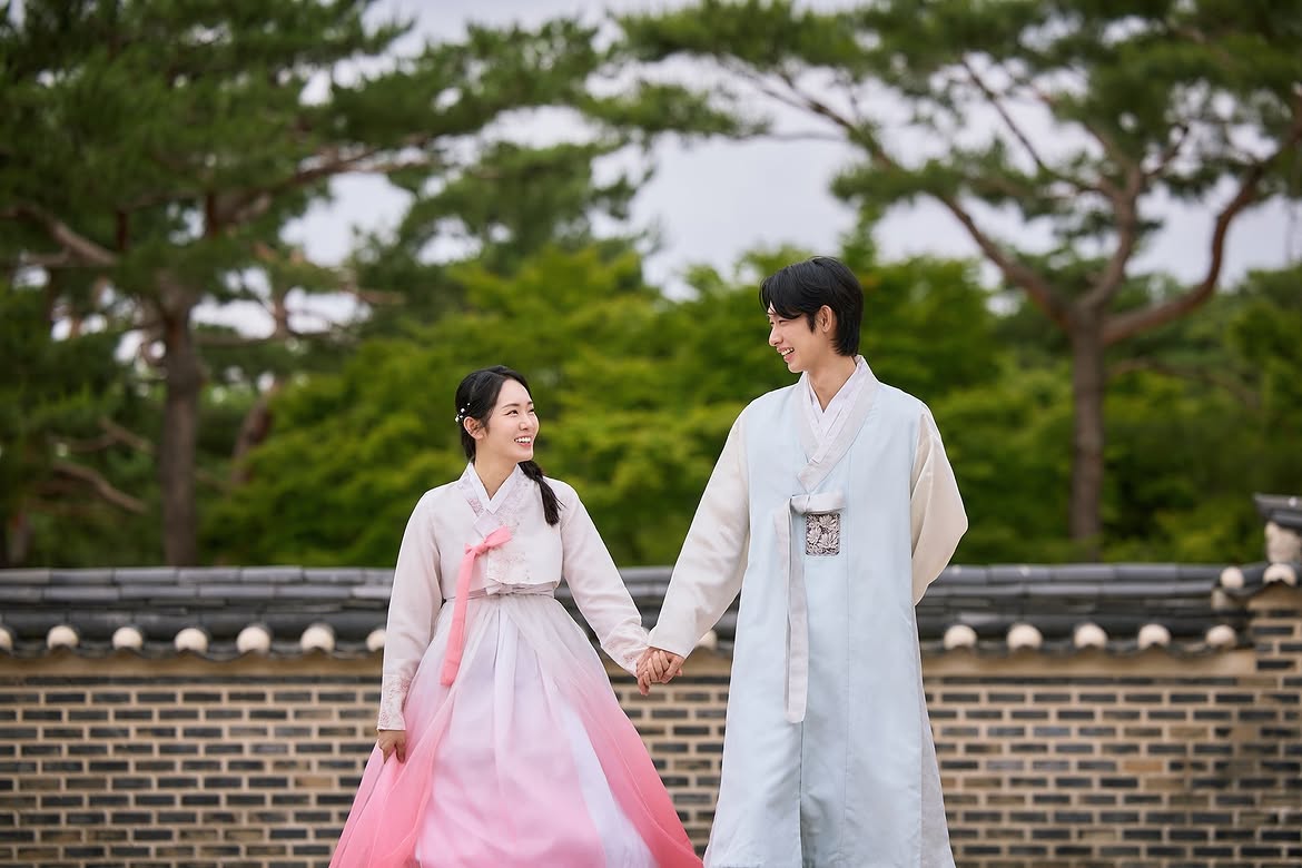 Couple in hanbok holding hands beside a brick wall